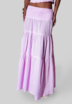 Kvinde iført et højtaljet, lagdelt lilafarvet maxinederdel med lodrette striber og en crop top med flæser mod en ensfarvet grå baggrund.