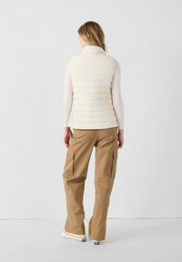 Gilet imbottito di colore crema chiaro con cuciture orizzontali e colletto alto, abbinato a pantaloni cargo beige con più tasche e vestibilità comoda.
