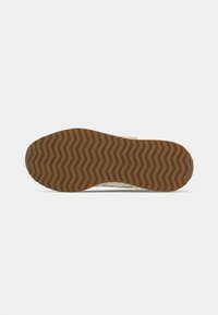 Turnschuhe mit einer strukturierten braunen Gummisohle, die ein Zickzackmuster aufweist. Das Obermaterial besteht aus einem hellen Material mit sichtbaren Nähten.