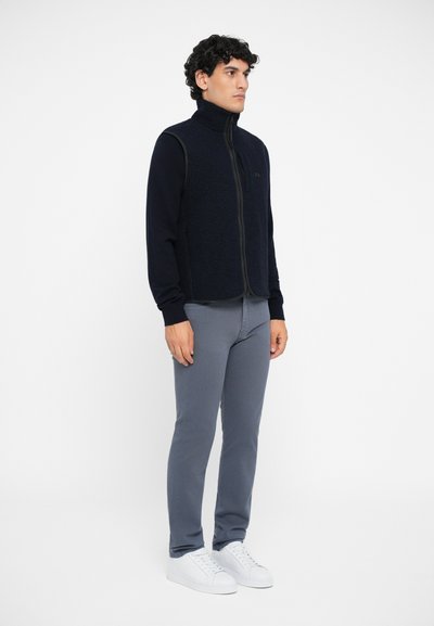 Marineblaue Fleece-Weste mit hohem Kragen und durchgehendem Reißverschluss, kombiniert mit grauen, körperbetonten Hosen und weißen Sneakers. Glatte Textur und minimalistisches Design.