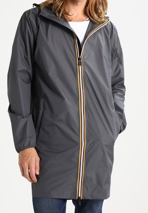 Personne portant un long imperméable gris foncé avec une fermeture éclair rayée et des poignets élastiques, les mains partiellement dans les poches, sur un jean bleu.