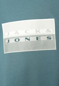 Μπλε βαμβακερό μπλουζάκι με λευκό ορθογώνιο λογότυπο με διάτρητη υφή και έντονα γράμματα "JACK & JONES" σε διάφορες αποχρώσεις του γκρι.