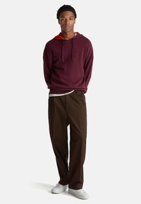 Sudadera con capucha color burdeos con interior rojo, de corte holgado, combinada con pantalones anchos marrones y zapatillas blancas, de pie contra un fondo sencillo.