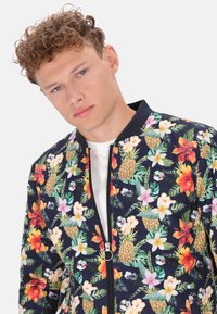 Giacca bomber floreale in blu scuro, caratterizzata da fiori vivaci e ananas. Chiusura con zip e tirante rotondo, colletto a costine. Tessuto leggero.