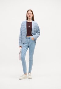 Svetlo modrý kardigán s vertikálnymi pruhmi, hnedý krátky tričko s logom, svetlo modré džínsy, biele tenisky a svetloružová taška.