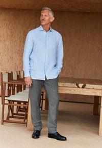 Lichtblauw linnen overhemd met knopen, grijze broek en zwarte schoenen met blauwe accenten. Staand naast een houten tafel in een neutrale omgeving.