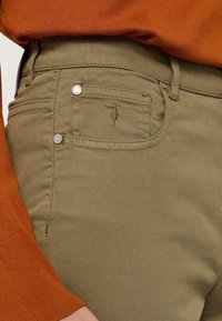 Olijfgroene broek met een getailleerd ontwerp, voorzien van een zak met decoratieve stikseldetails en accenten in zilverkleurig metaal.