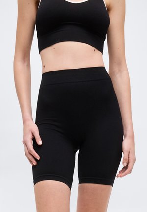 Frau trägt ein schwarzes, bauchfreies Tanktop und hoch taillierte schwarze Biker-Shorts, steht mit entspannten Händen seitlich am Körper.