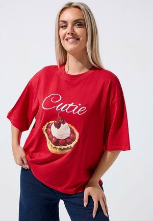 Smilende kvinde med langt blondt hår iført en rød oversize T-shirt med teksten "Cutie" og grafik af en kirsebærtærte, poserer med hænderne på hofterne.