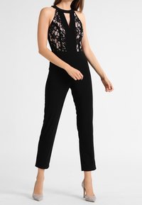 Schwarzer Jumpsuit mit Spitzentop, Trägern im Halterneck-Stil und Ausschnittdetails. Eng anliegende Beine und strukturierte Spitzenelemente verleihen visuelles Interesse. Neutrale Absätze.