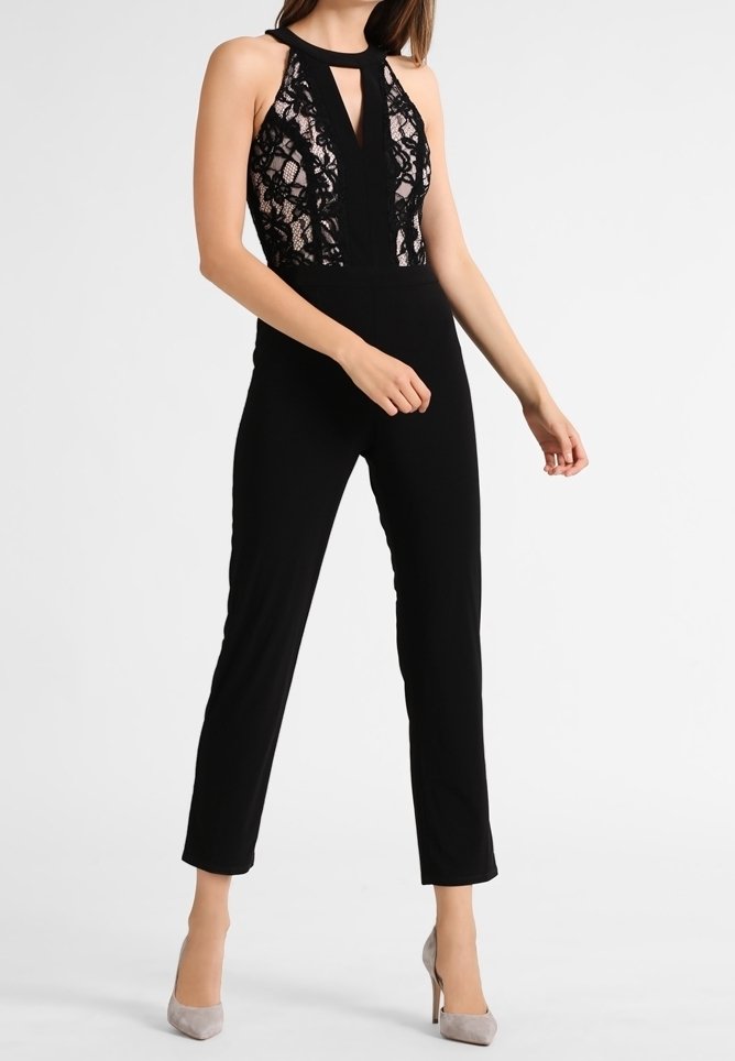 Schwarzer Jumpsuit mit Spitzentop, Trägern im Halterneck-Stil und Ausschnittdetails. Eng anliegende Beine und strukturierte Spitzenelemente verleihen visuelles Interesse. Neutrale Absätze.