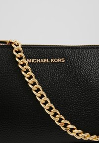 Bolso de cuero negro con un acabado texturizado, que presenta una correa de cadena dorada y el logo "MICHAEL KORS" en letras doradas. Cierre de cremallera en la parte superior.