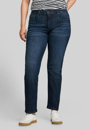 Persoon draagt donkerblauwe jeans met rechte pijpen, witte sneakers met rubberen zolen en een zwart-wit gestreept shirt tegen een grijze achtergrond.