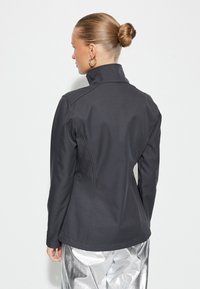 Chaqueta negra ajustada con cuello alto y diseño sin costuras; fabricada con tejido suave y ligero. La vista trasera muestra una silueta entallada.