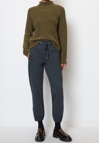 Olivgrüner gestrickter Pullover mit hohem Kragen, kombiniert mit dunkelgrauen tapered Jogginghosen mit elastischen Bündchen und verstellbarem Bund. Schwarze Schuhe.