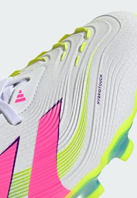 Hvit fotballstøvel med teksturert overdel, rosa og gule detaljer, med en "HYBRIDTOUCH"-etikett. Designet for ytelse med en strømlinjeformet fasong.