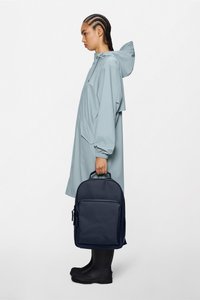 Grauer wasserdichter Regenmantel mit Kapuze, getragen über schwarzen Gummistiefeln, hält einen marineblauen Rucksack mit einer Vordertasche und einem oberen Griff.