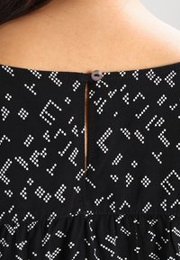 Czarna sukienka z tkaniny w rozproszony wzór białych kropek, z wycięciem w kształcie kluczyka z tyłu i okrągłym zapięciem na guzik.