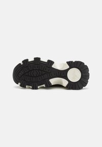 Fekete-fehér sneaker talp, amely robusztus dizájnnal rendelkezik, texturált gumival a tapadás érdekében, és hangsúlyos barázdázott mintázattal, kör alakú logó részlettel.