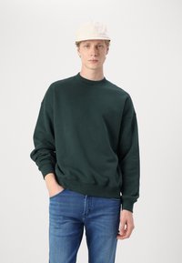 Sudadera verde oscura con cuello redondo, corte holgado, puños acanalados, llevada sobre jeans azul claro, complementada con una gorra beis.