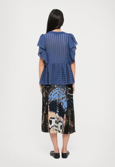 Woman with long dark hair wears a blue striped ruffle-sleeve blouse and a pleated skirt with a large colorful abstract face design, facing away.