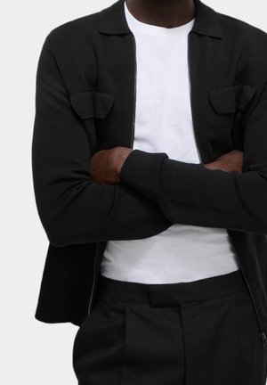 Persona con le braccia incrociate che indossa una giacca nera con cerniera sopra una camicia bianca e pantaloni neri, mostrata dalle spalle alla vita.