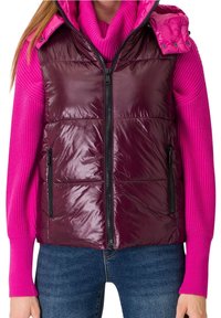 Femme portant une doudoune sans manches brillante de couleur violet foncé sur un pull côtelé rose vif et un jean bleu, la doudoune est zippée et les poches sont visibles.