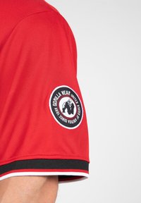 Rode mesh sportshirt met zwart-witte gestreepte accenten. Heeft een geborduurd rond logo op de mouw.