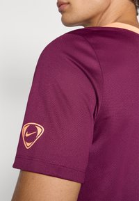 Camiseta atlética bordeau com um tecido texturizado. Apresenta mangas curtas e um logo da Nike em laranja claro na manga esquerda.
