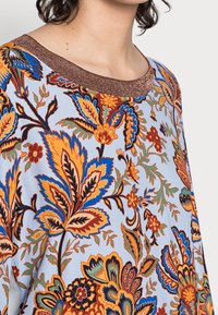 Blouse bleue à motifs florauxintriqués en orange et vert, avec un col en tricot brun brillant et une texture douce.