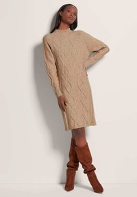 Beige kabelgebreide trui jurk met lange mouwen, een hoge halslijn en ribgebreide manchetten. Het model draagt een paar bruine kniehoge slouch laarzen.