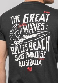T-shirt en coton gris foncé avec un texte audacieux en blanc et rouge. Le graphisme inclut une vague et une planche de surf avec des phrases sur les vagues et une plage en Australie.