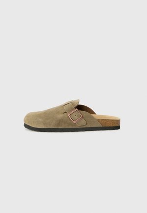 Brauner Wildleder-Slipper mit verstellbarem Schnallenriemen und schwarzer Sohle vor einfarbigem grauem Hintergrund.