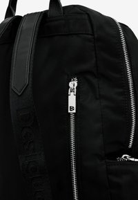 Zwarte rugzak gemaakt van nylon met een gestructureerd oppervlak, voorzien van zilveren ritsen en een geweven riem met het woord "Design" erin geperst.