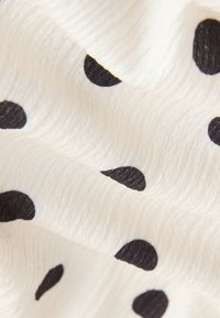 Fehér, texturált szövet szabálytalan fekete pöttyökkel, közeli képen, látható redőkkel és gyűrődésekkel.