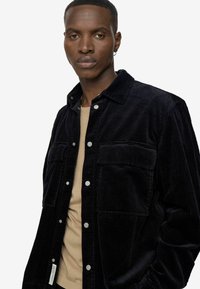 Black corduroy button-up shirt with two chest pockets, silver hardware, and a textured surface, layered over a beige t-shirt.
