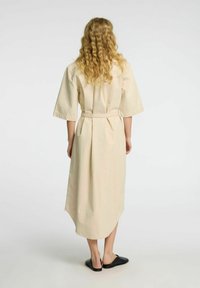 Beige bomulds midi kjole med korte ærmer, indsnævret talje og buet kant. Båret med sorte slip-on sko, vist fra ryggen.