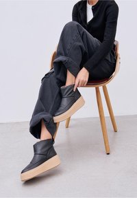 Persona sentada en una silla de madera, vestida con pantalones negros y un suéter, ajustándose las zapatillas altas negras con suelas beige.