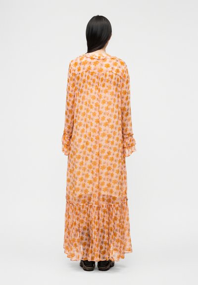 Γυναίκα με μακριά σκούρα μαλλιά, φορώντας μακρύ διαφανές φόρεμα ροδακινί με εκτύπωση πορτοκαλί λουλουδιών, που στέκεται με την πλάτη, με ορατά μαύρα παπούτσια.