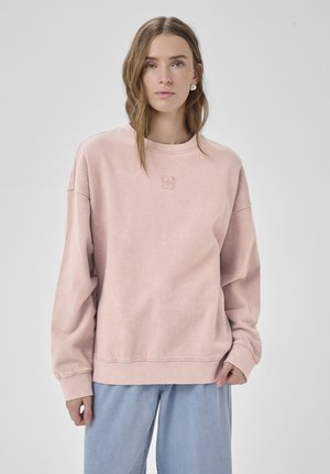 Valgepruunide juustega naine kannab üle suurust kahvaturoosat pusad ja helesiniseid teksaseid, seisab halli ühevärvilise tausta ees.