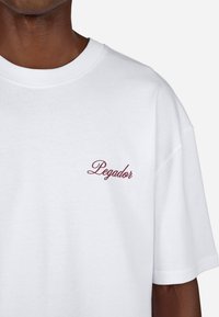 Biała bawełniana koszulka z okrągłym dekoltem, z bordowym haftowanym logo "Pegador" na lewej piersi. Luźny krój z krótkimi rękawami.