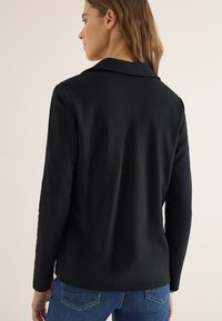 Femme portant un blazer noir à manches longues avec un col, vue de dos, associé à un jean en denim bleu.