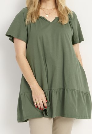 Woman wearing loose olive green short-sleeve dress with ruffled hem, beige pants, gold rings, and red nail polish, standing against white background.