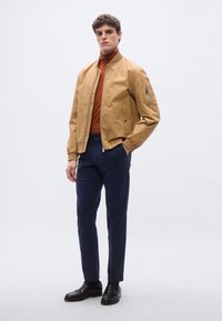 Beige bomberjacka med ribbad krage, brun polotröja, marinblå byxor och svarta loafers. Modellen står mot en ljusgrå bakgrund.