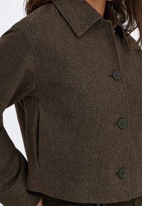 Veste marron texturée à boutons avec un large col portée par une personne ayant la main posée sur la hanche.