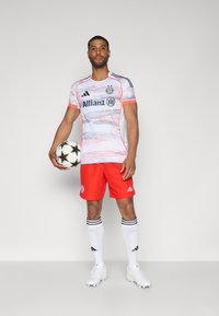 La divisa del Bayern Monaco presenta una maglietta bianca e rosa con motivo, pantaloni rossi, calzini bianchi con strisce nere e un pallone da calcio bianco e nero.