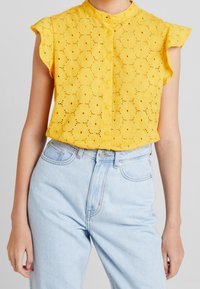 Yellow eyelet blouse with short flutter sleeves and a button-up front, paired with light blue high-waisted denim jeans.