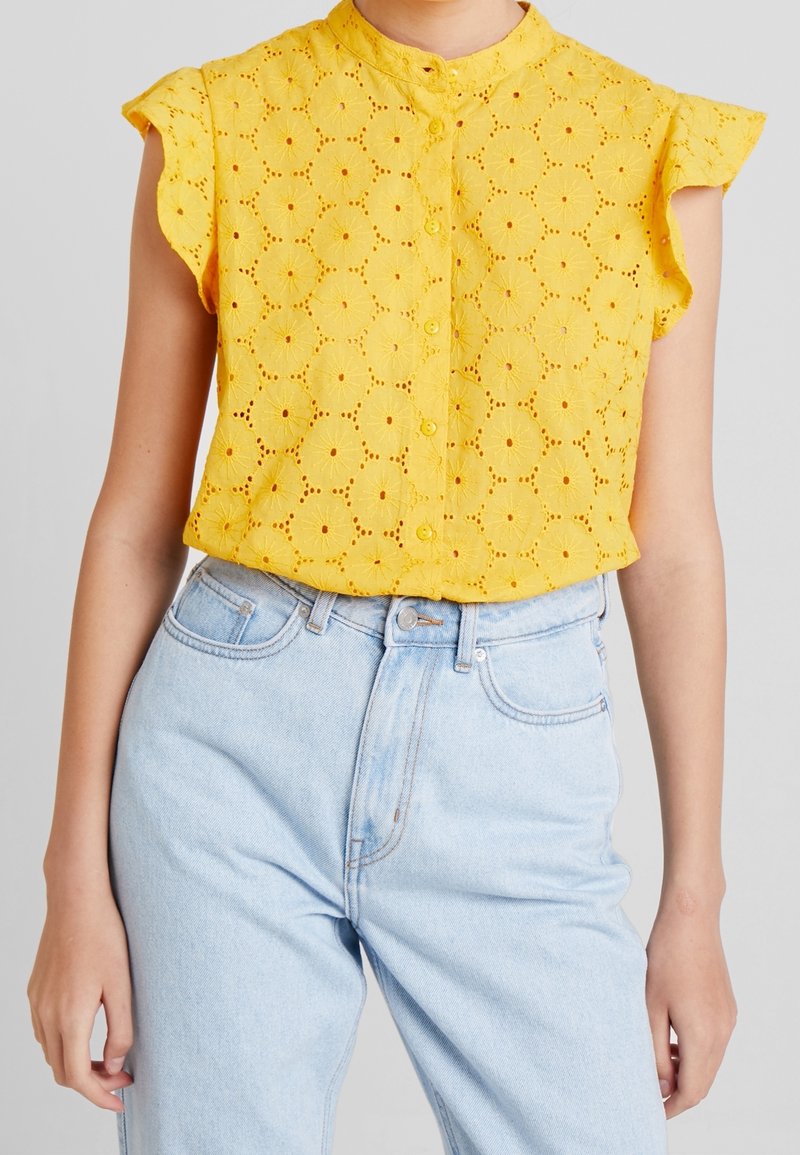 Yellow eyelet blouse with short flutter sleeves and a button-up front, paired with light blue high-waisted denim jeans.