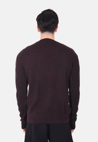 Maglione marrone lavorato a maglia con una texture a coste, maniche lunghe e scollo rotondo, visto da dietro, abbinato a pantaloni scuri.