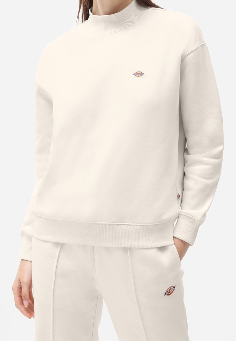 Sudadera de color beige claro con un cuello alto, puños acanalados y dobladillo. Presenta un pequeño logo bordado en el pecho y una textura suave.